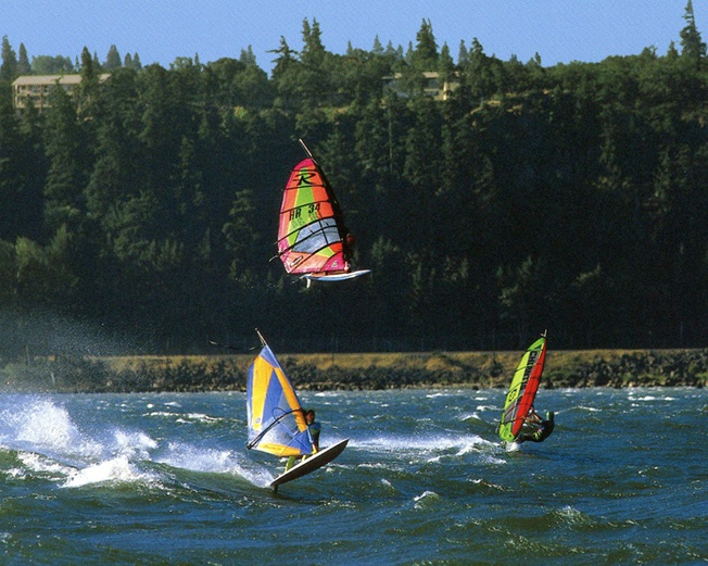 three people windsurfing, one airborne, cruise the river winds