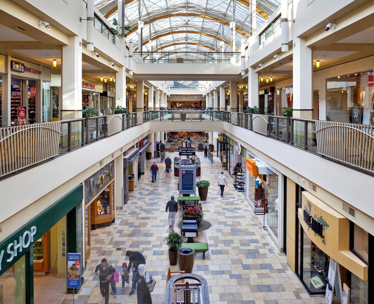 shoppers explore an indoor three-story shopping mall lined with storefronts and illuminated by natural light from above