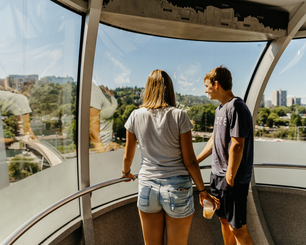 Two people stand inside a rounded aerial tram cabin, observing the forest and buildings below