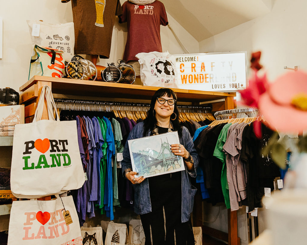Co-founder Cathy Zwicker holds up a print of a Portland bridge, surrounded by tote bags that say "Portland" and other Portland-related merch.