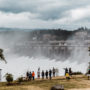People stnad on a river bank observing water flowing over a large dam