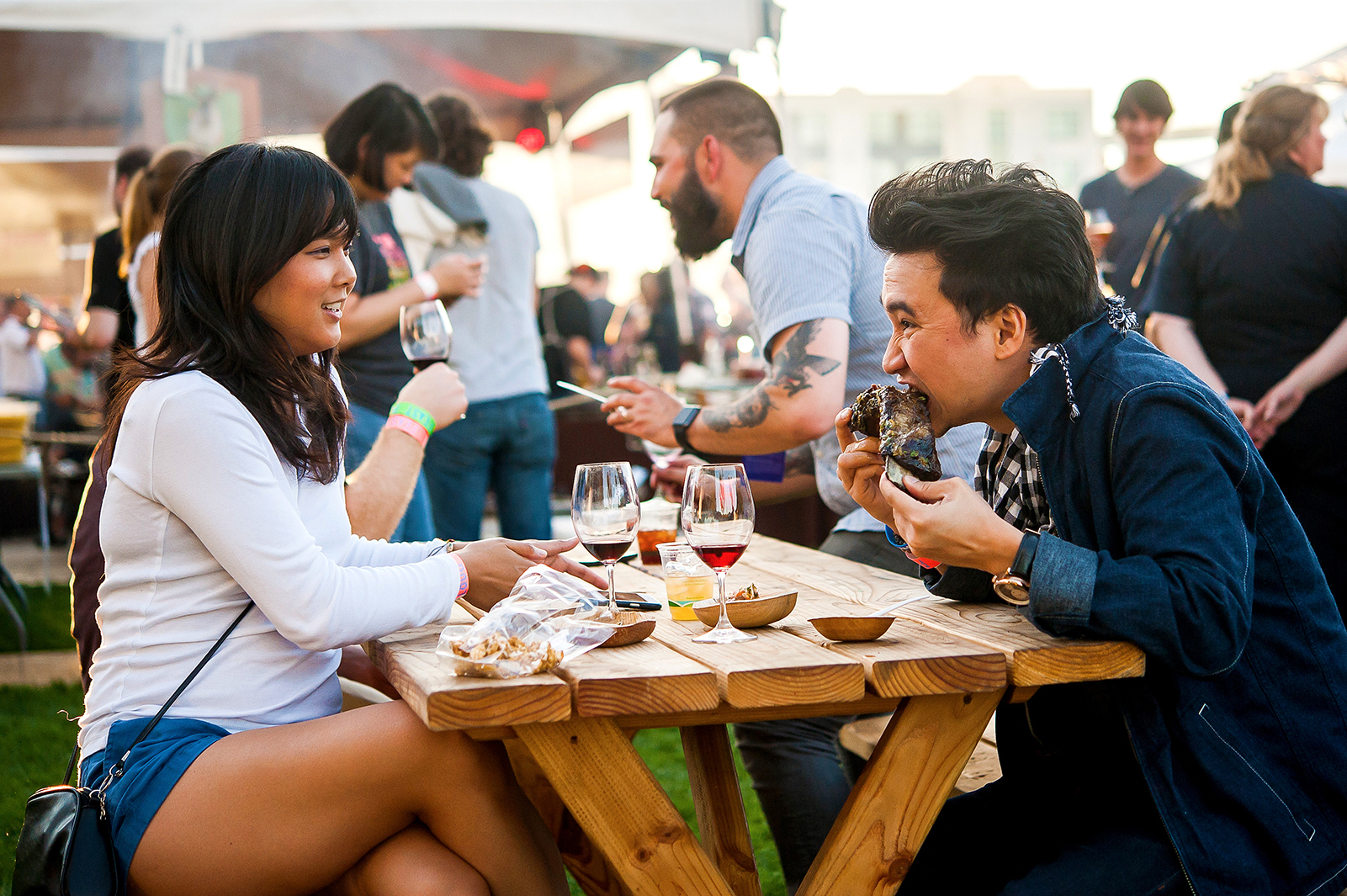 two people eating food and drinking wine at a picnic table during a busy food festival