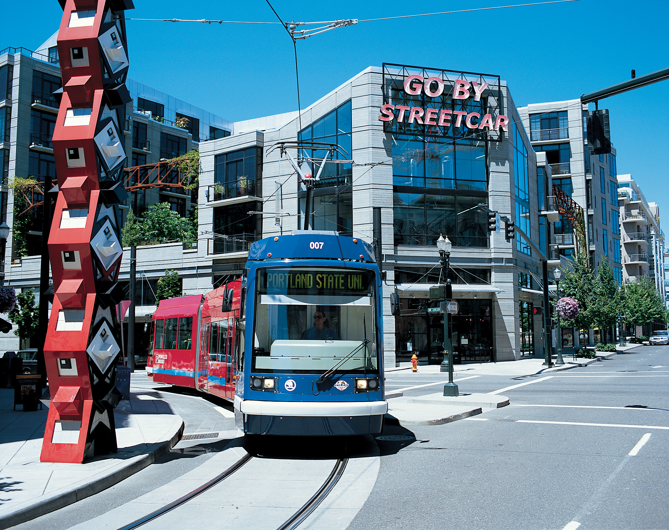 A streetcar passes by a large, red sculpture in front of a building with a neon sign reading, \"Go By Streetcar.\"