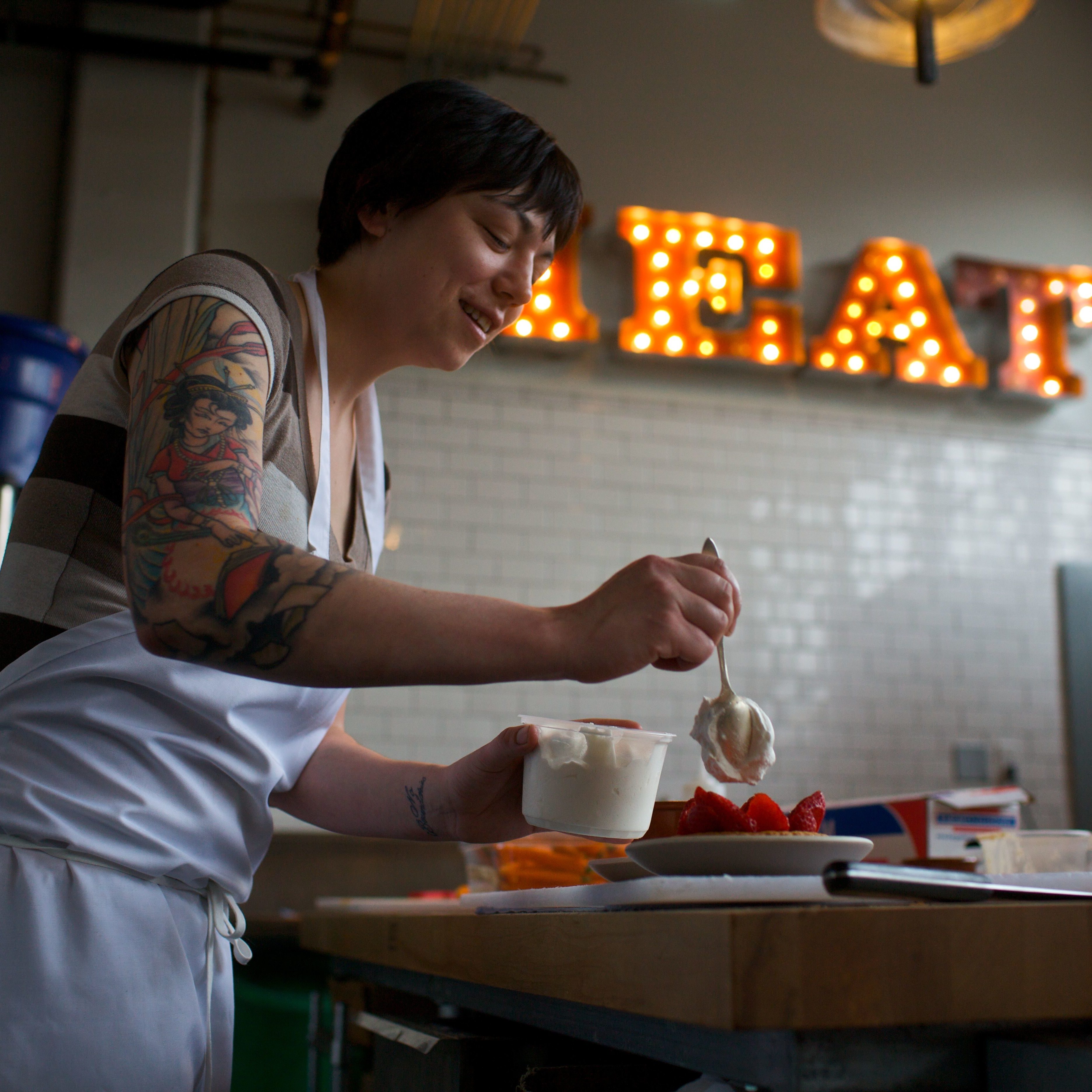 A chef tops a strawberry dessert with whipped cream in front of a neon sign reading "MEAT"