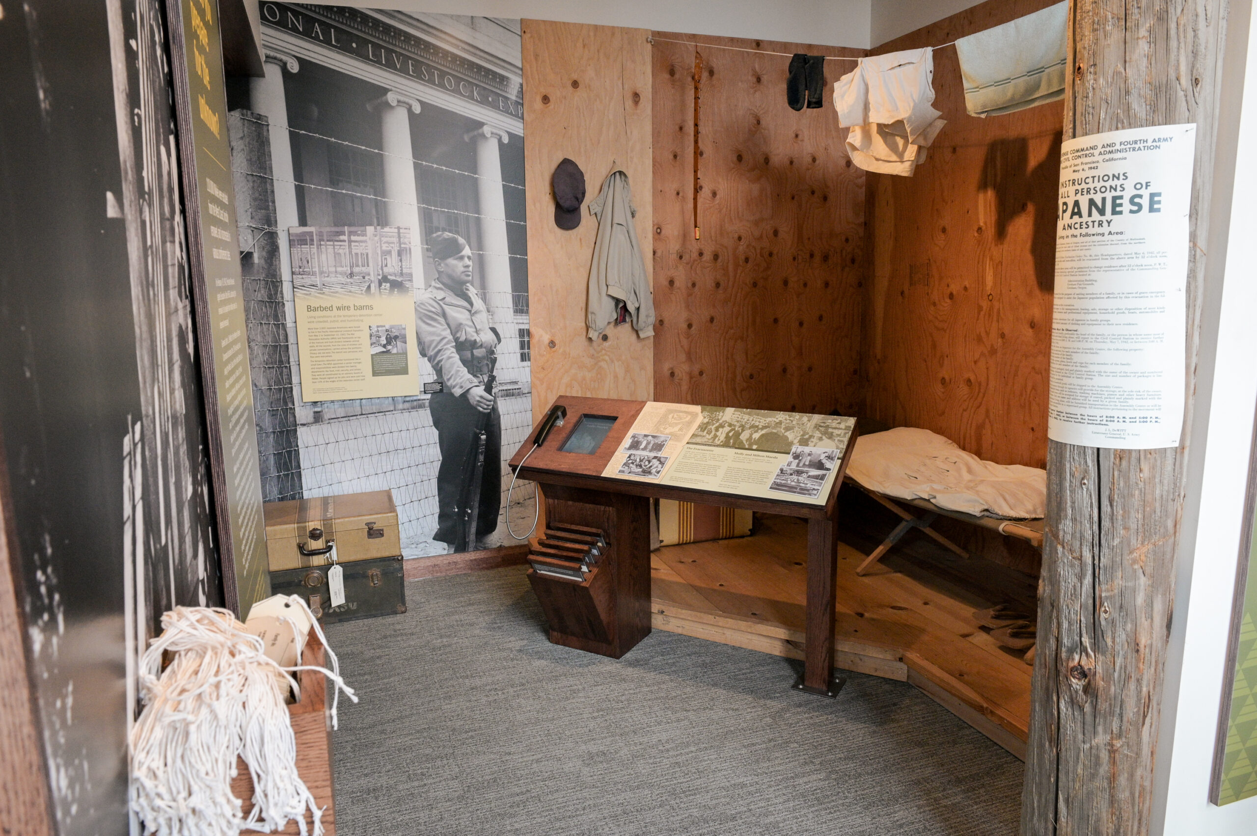small living space including a cot and hung up laundry with image of a barbed wire fence surrounding, depicting Japanese internment living quarters