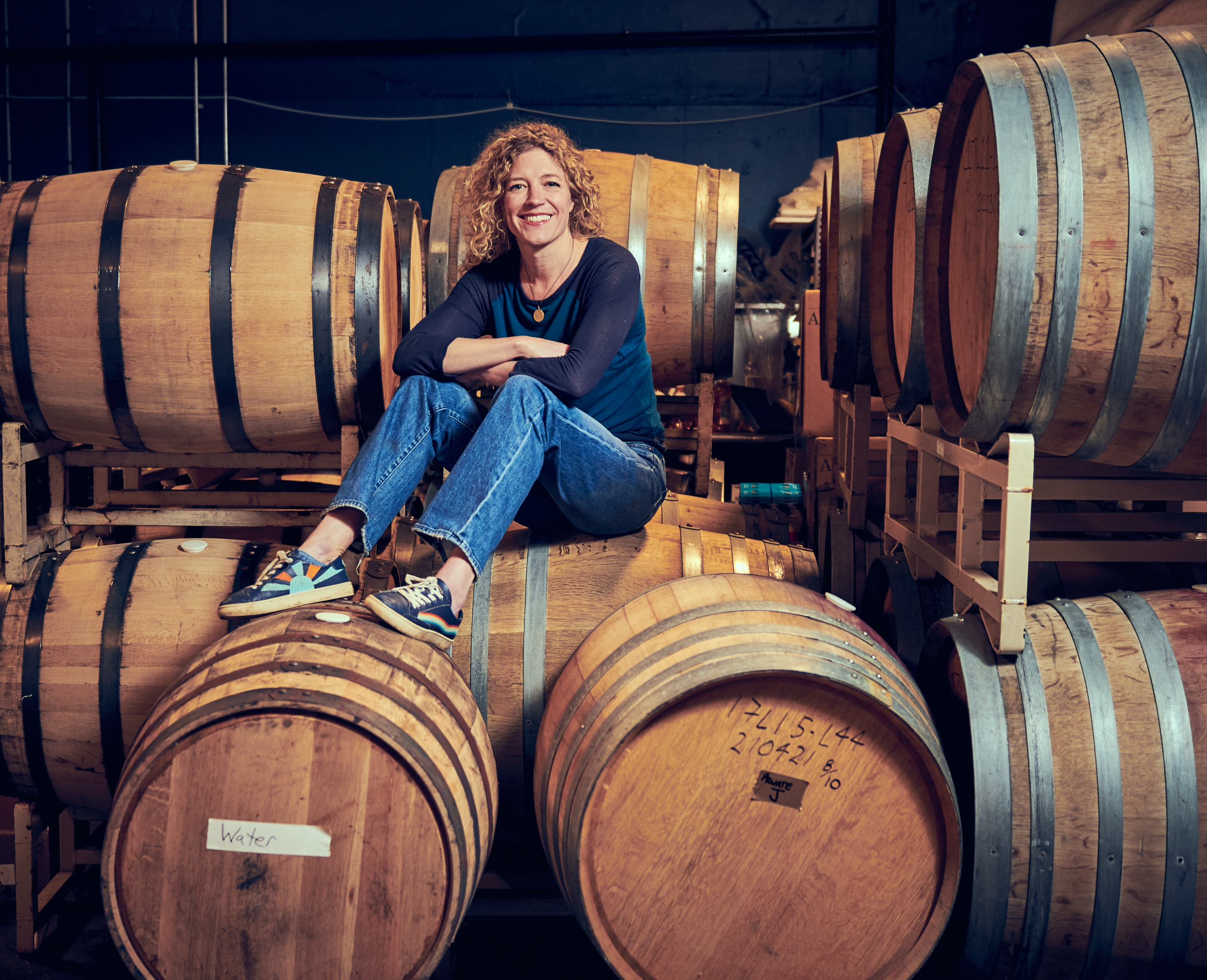 A woman in long-sleeved t-shirt, jeans and sneakers sits on top of large wooden barrels