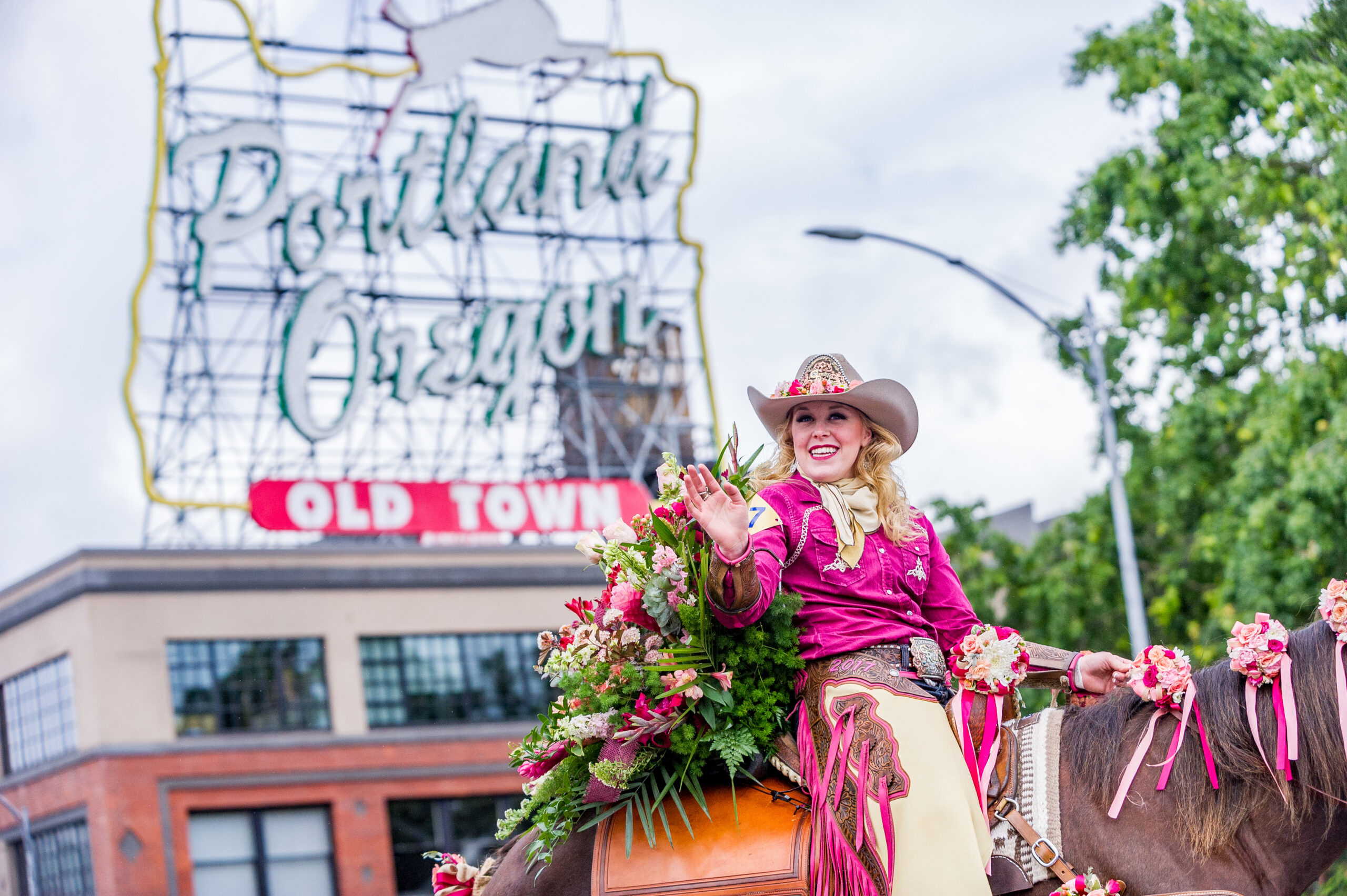 woman in hat and chaps ride a rose decorated horse in a parade with Portland's white stag sign in the background