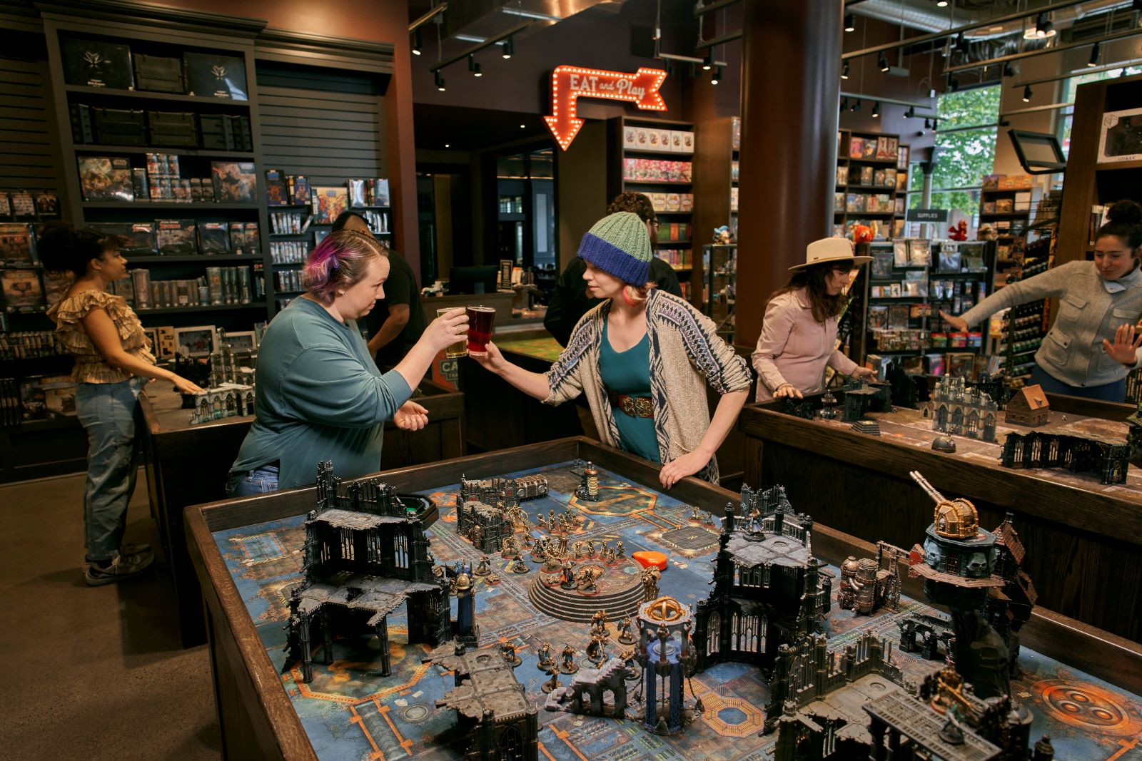 Two friends clinking glasses over a large tabletop board game.