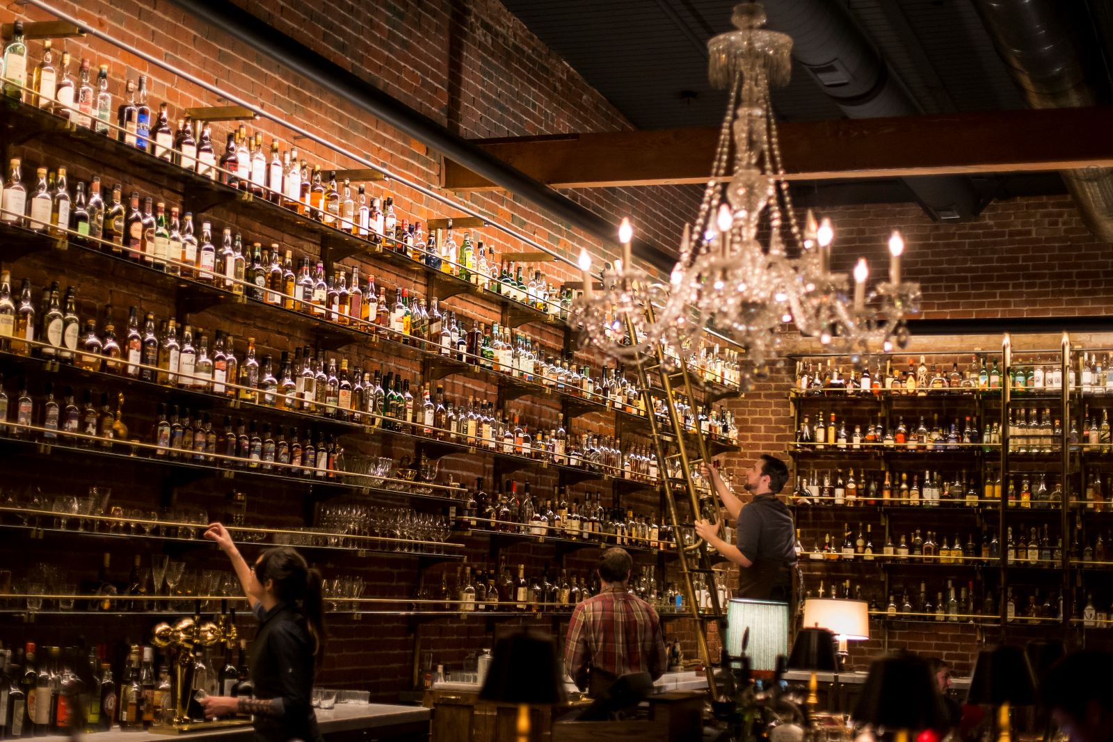 several bartenders peruse shelves housing a vast collection of whiskey bottles