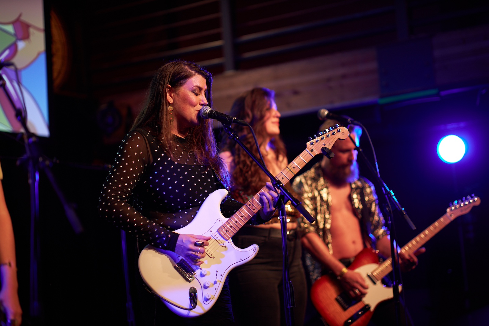person sings in to a microphone while playing a guitar with two band members behind on a stage.