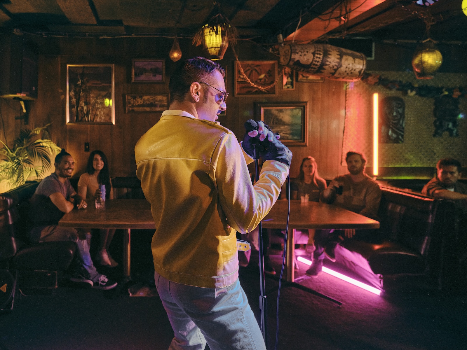 a person in a yellow leather-looking jacket singing in to a microphone in front of patrons in a bar