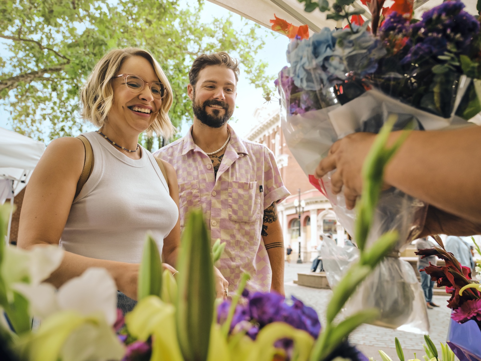 people smiling on a sunny day in front of bunches of flowers while someone off screen is handing them a bundle of cut flowers in a plastic bag
